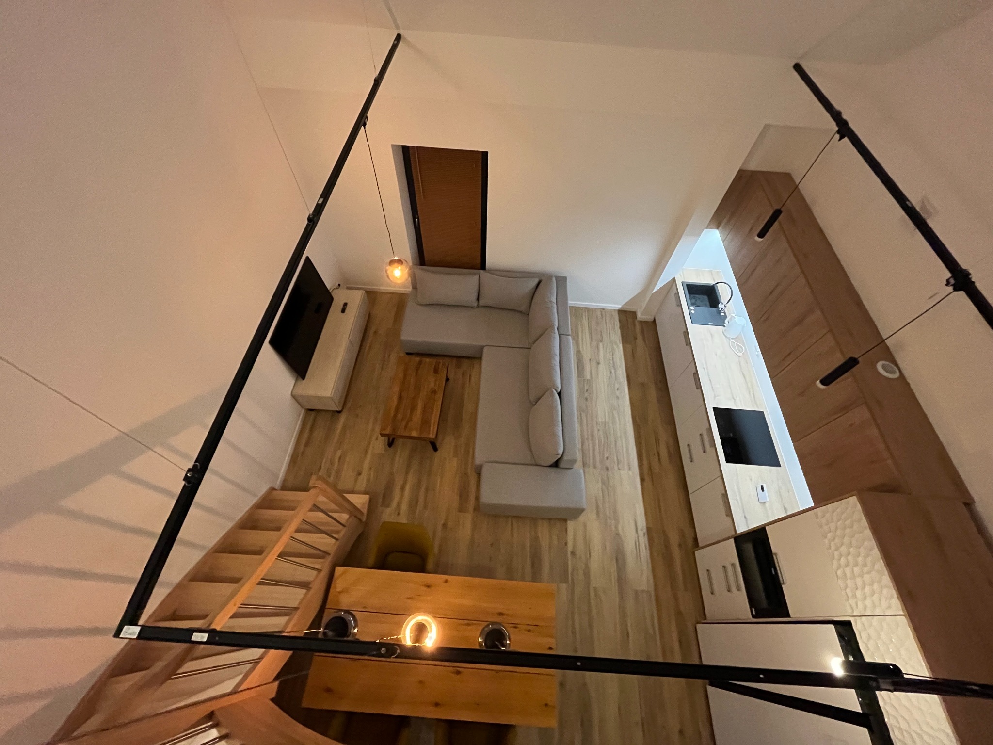 Wnętrze mieszkania z antresolą, widok z góry. Jasne drewno na podłodze, szara sofa narożna, drewniany stół. Kuchnia z białymi szafkami i drewnianymi akcentami. Schody na antresolę.