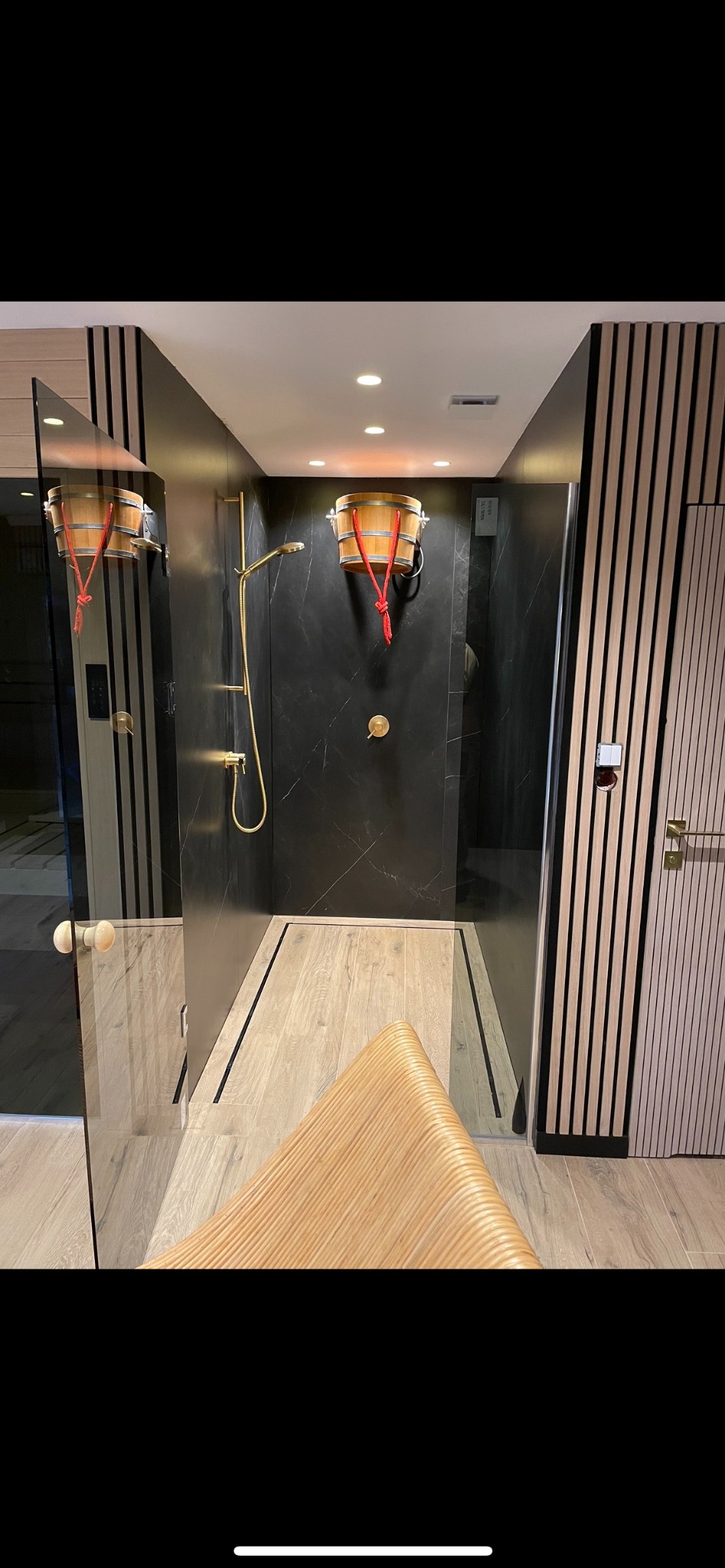 Nowoczesna kabina prysznicowa z drewnianymi akcentami i złotą armaturą we Wrocławiu. Czarna ściana z marmurowym wzorem, drewniana podłoga i siedzisko.