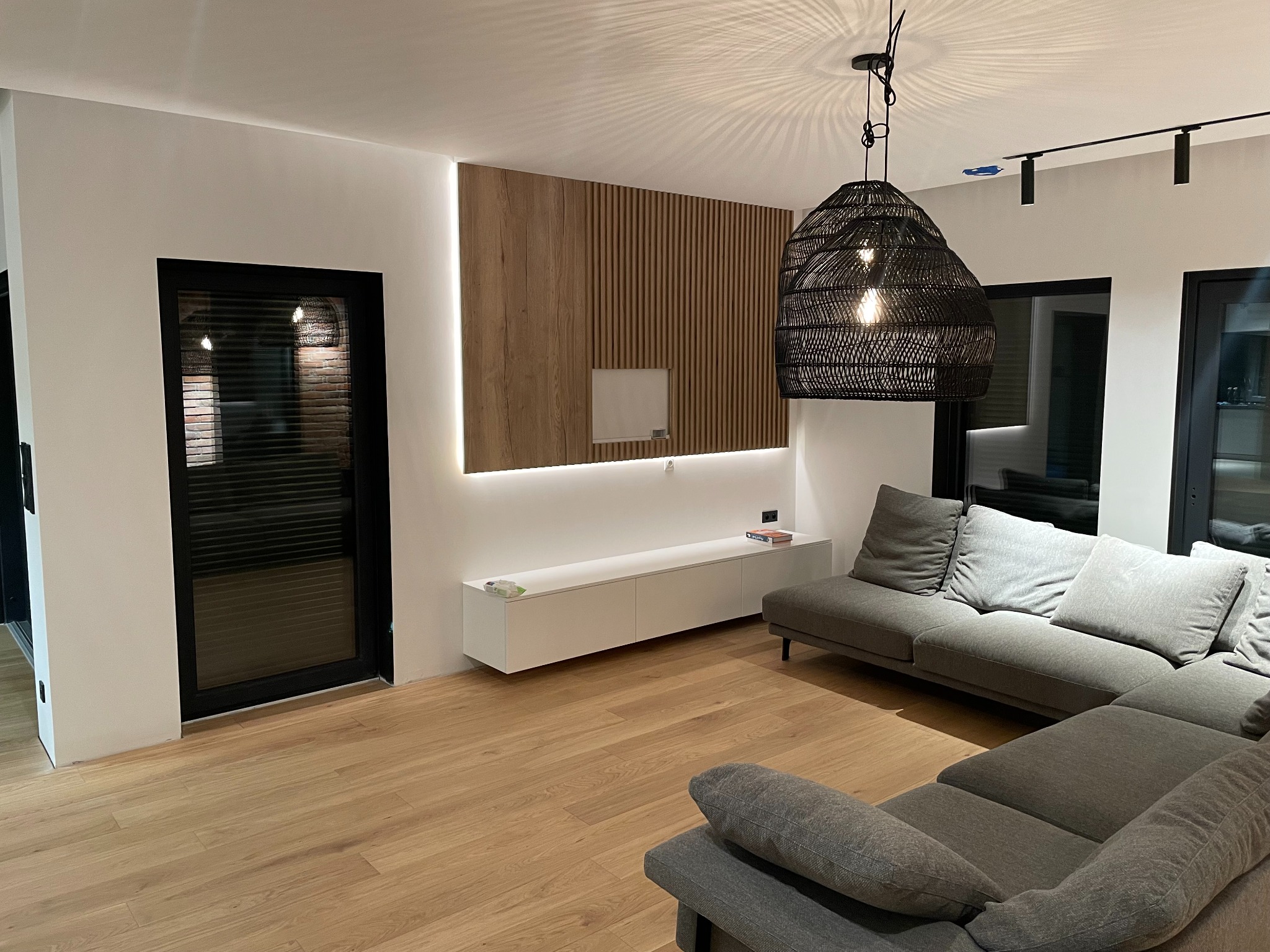 Nowoczesny salon po remoncie: drewniana ściana z podświetleniem LED, szara sofa narożna, parkiet i czarne ramy drzwi. W tle widoczna lampa wisząca z abażurem z naturalnego materiału.
