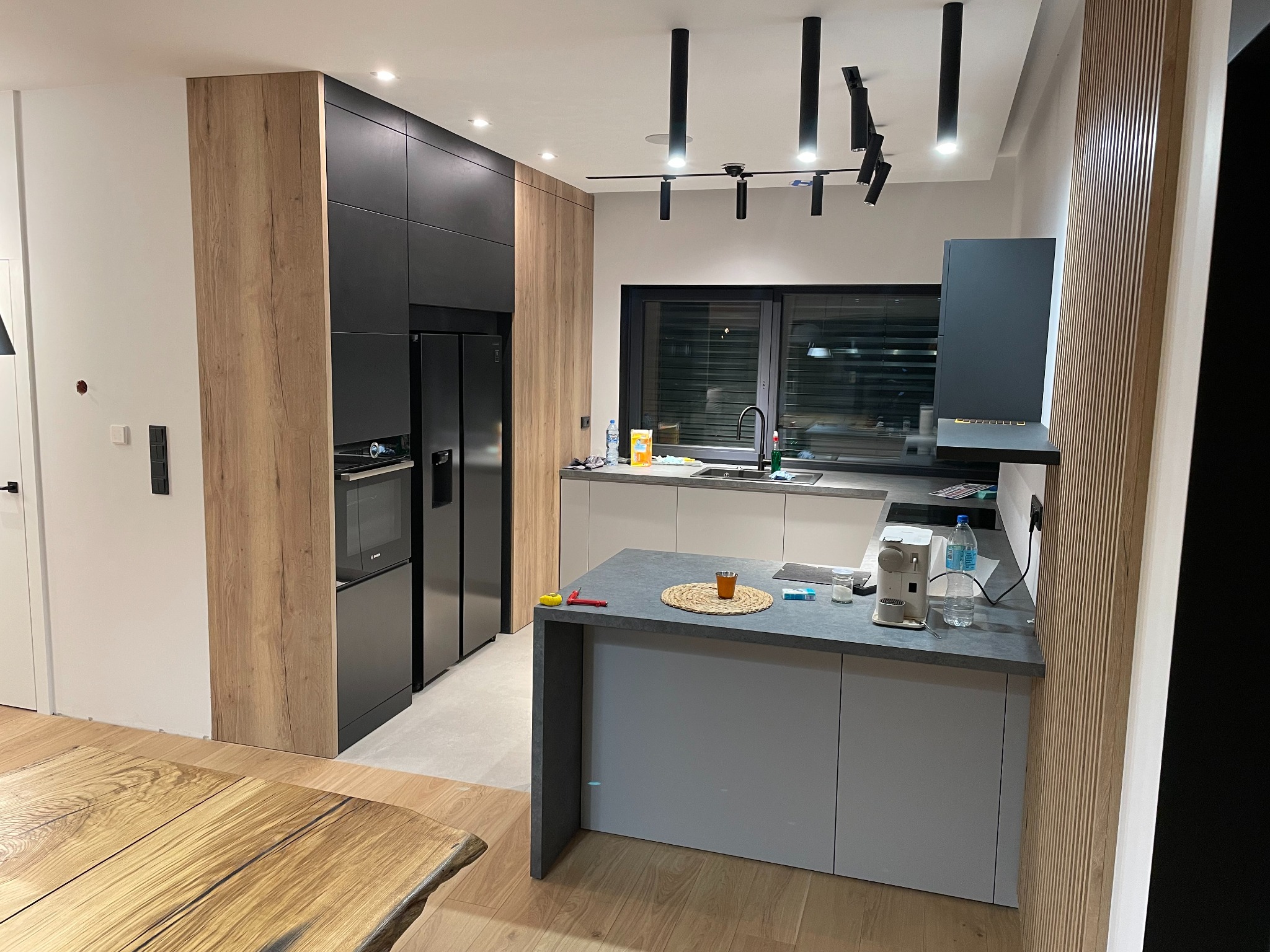 Nowoczesna kuchnia z wyspą, czarnymi szafkami i drewnianymi akcentami. Widoczne oświetlenie punktowe i okno. Na blacie akcesoria kuchenne i napoje.
