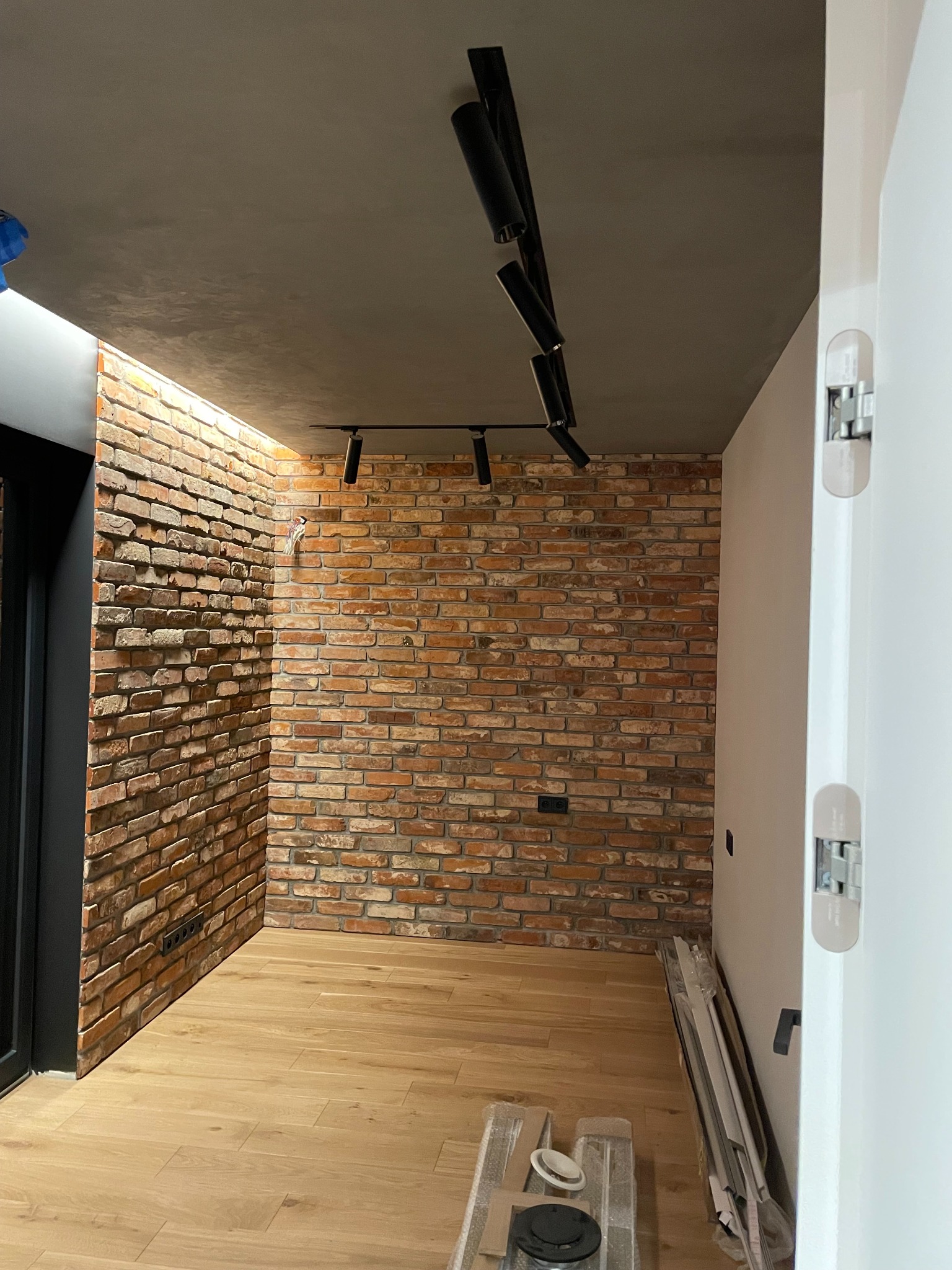 Wykończone wnętrze z ceglaną ścianą, drewnianą podłogą i nowoczesnym oświetleniem sufitowym. Widoczne elementy instalacji elektrycznej i wykończeniowe.