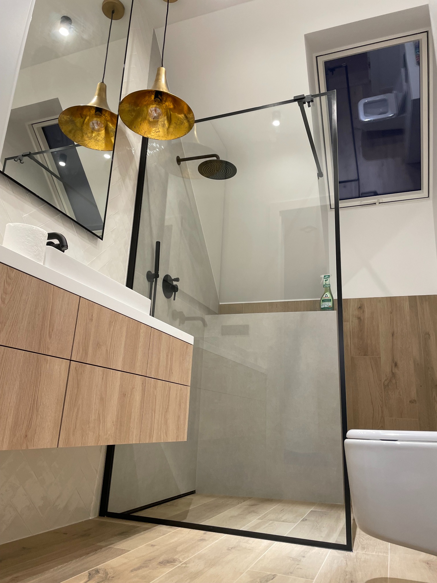Nowoczesna łazienka z kabiną prysznicową typu walk-in, drewnianą szafką pod umywalką i złotymi lampami wiszącymi. Widoczne okno i toaleta.