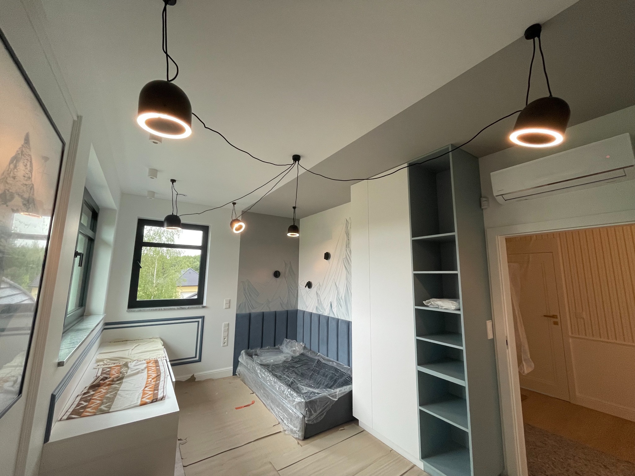 Pokój po remoncie: nowoczesne oświetlenie, tapicerowane wezgłowie łóżka, zabudowa meblowa. Widok na okno i inne pomieszczenie. Podłoga zabezpieczona folią.