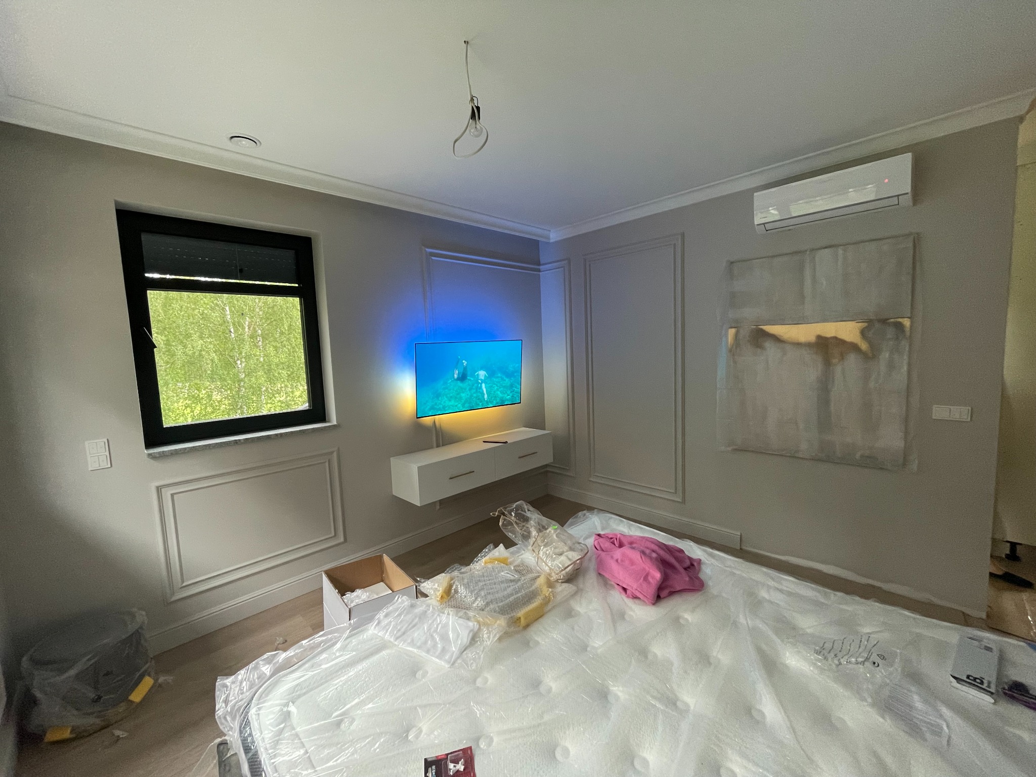 Pokój w trakcie wykańczania: szare ściany z dekoracyjnymi panelami, telewizor na ścianie z podświetleniem, klimatyzator, okno z widokiem na zieleń, materac zabezpieczony folią.