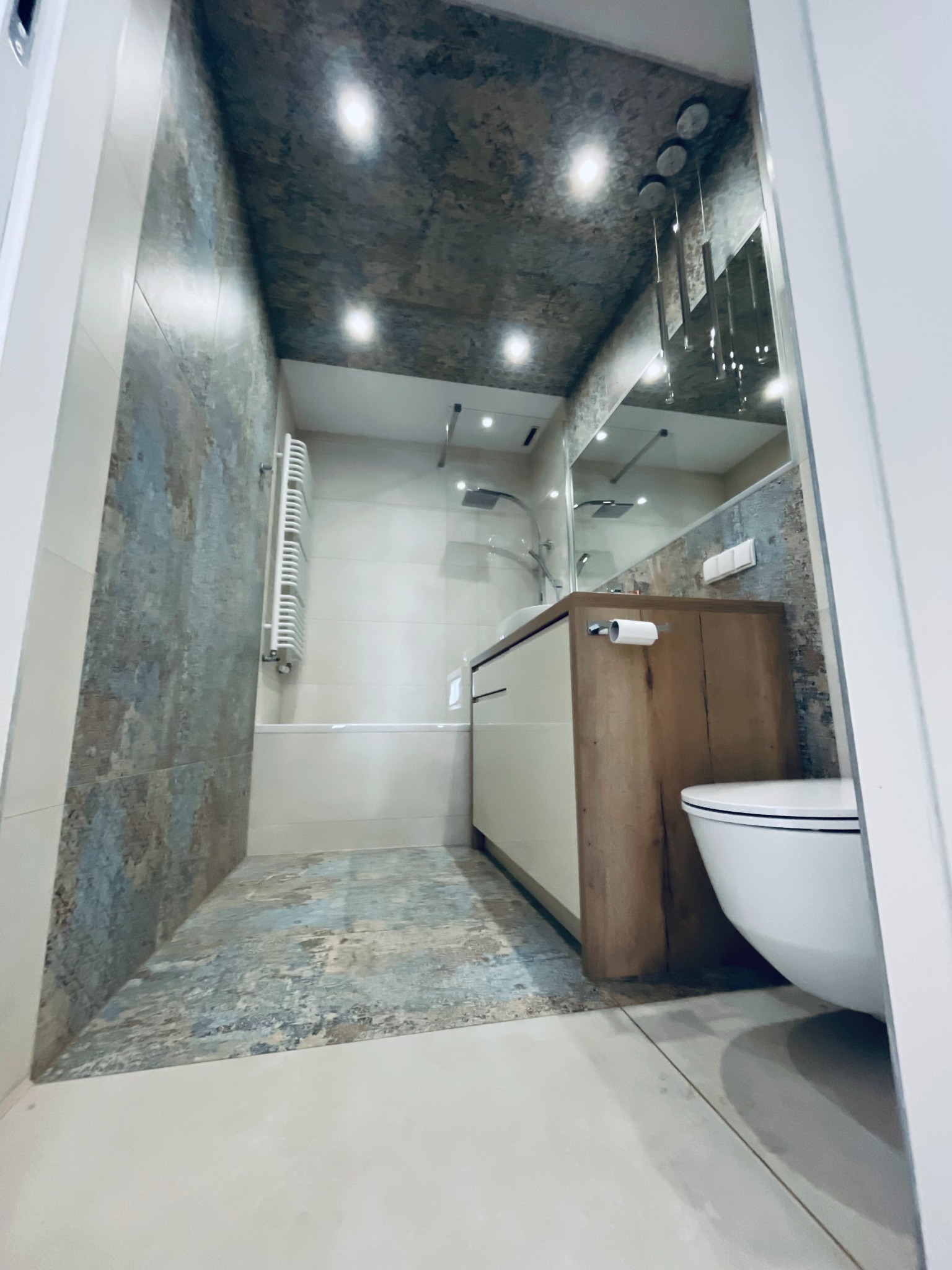 Nowoczesna łazienka z płytkami imitującymi kamień na ścianach i podłodze, białą armaturą i drewnianą szafką pod umywalką. Oświetlenie punktowe na suficie.