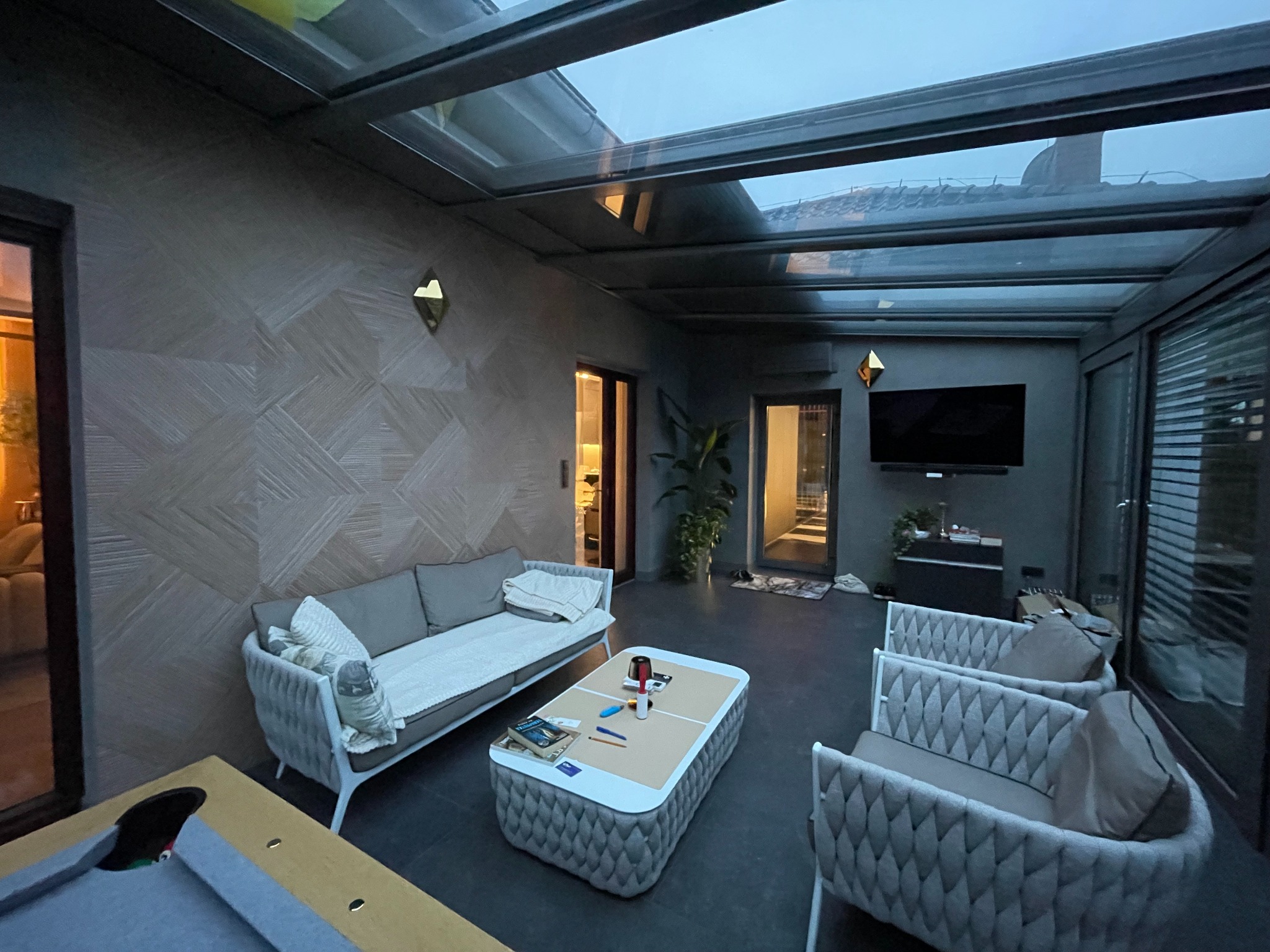 Zadaszony taras z meblami wypoczynkowymi: sofa, fotele i stolik kawowy. Ściana z dekoracyjną tapetą. Przeszklony dach z widokiem na niebo. Nowoczesny design.