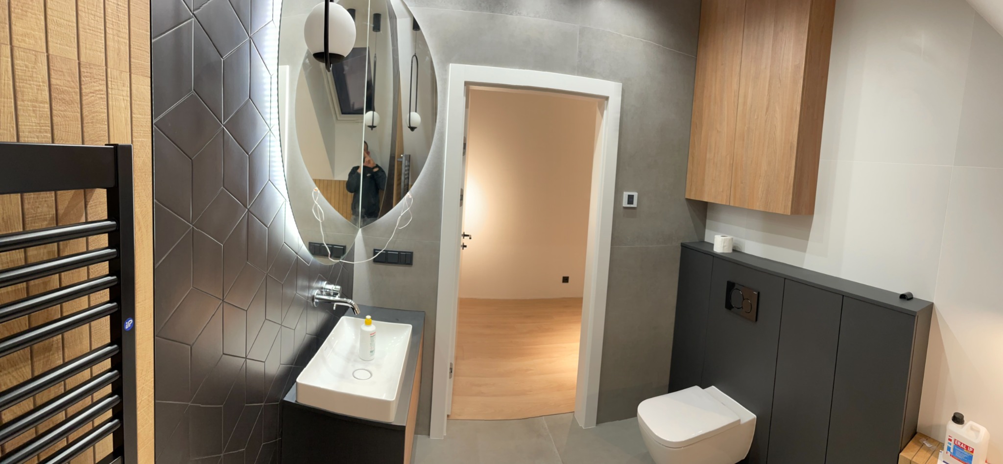 Nowoczesna łazienka po remoncie: geometryczne płytki, owalne lustro z podświetleniem LED, drewniane akcenty i wisząca toaleta. Widok na otwarte drzwi do innego pomieszczenia.