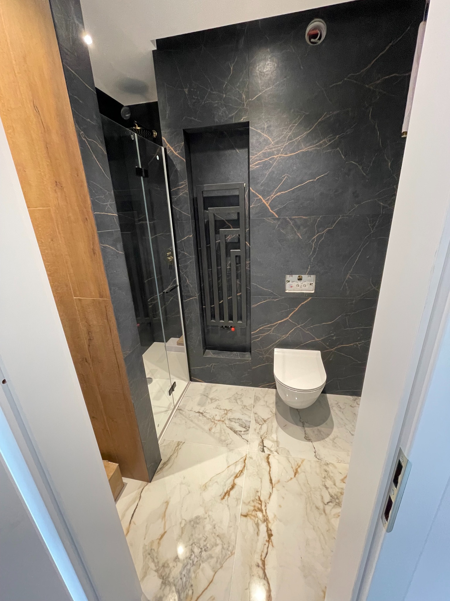 Nowoczesna łazienka z czarnymi płytkami imitującymi marmur z złotymi żyłami, białą armaturą i kabiną prysznicową. Widok zza otwartych drzwi.