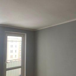 FP CONSTRUCTION - Jasnoszary pokój po malowaniu z białym sufitem i listwą. Widoczne okno balkonowe z widokiem na budynek. Fragment instalacji wystaje z sufitu.