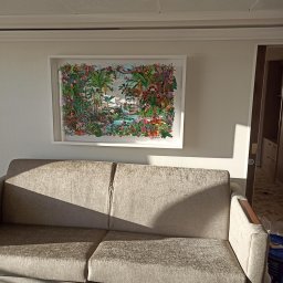 FP CONSTRUCTION - Salon z beżową sofą pod ścianą, na której wisi oprawiony w ramę obraz z motywem tropikalnym, oświetlony popołudniowym słońcem wpadającym przez okno.