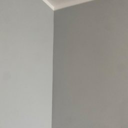 FP CONSTRUCTION - Świeżo pomalowany róg pokoju w odcieniach szarości, z widoczną białą listwą sufitową. Minimalistyczne ujęcie, ukazujące gładką powierzchnię ścian po malowaniu.
