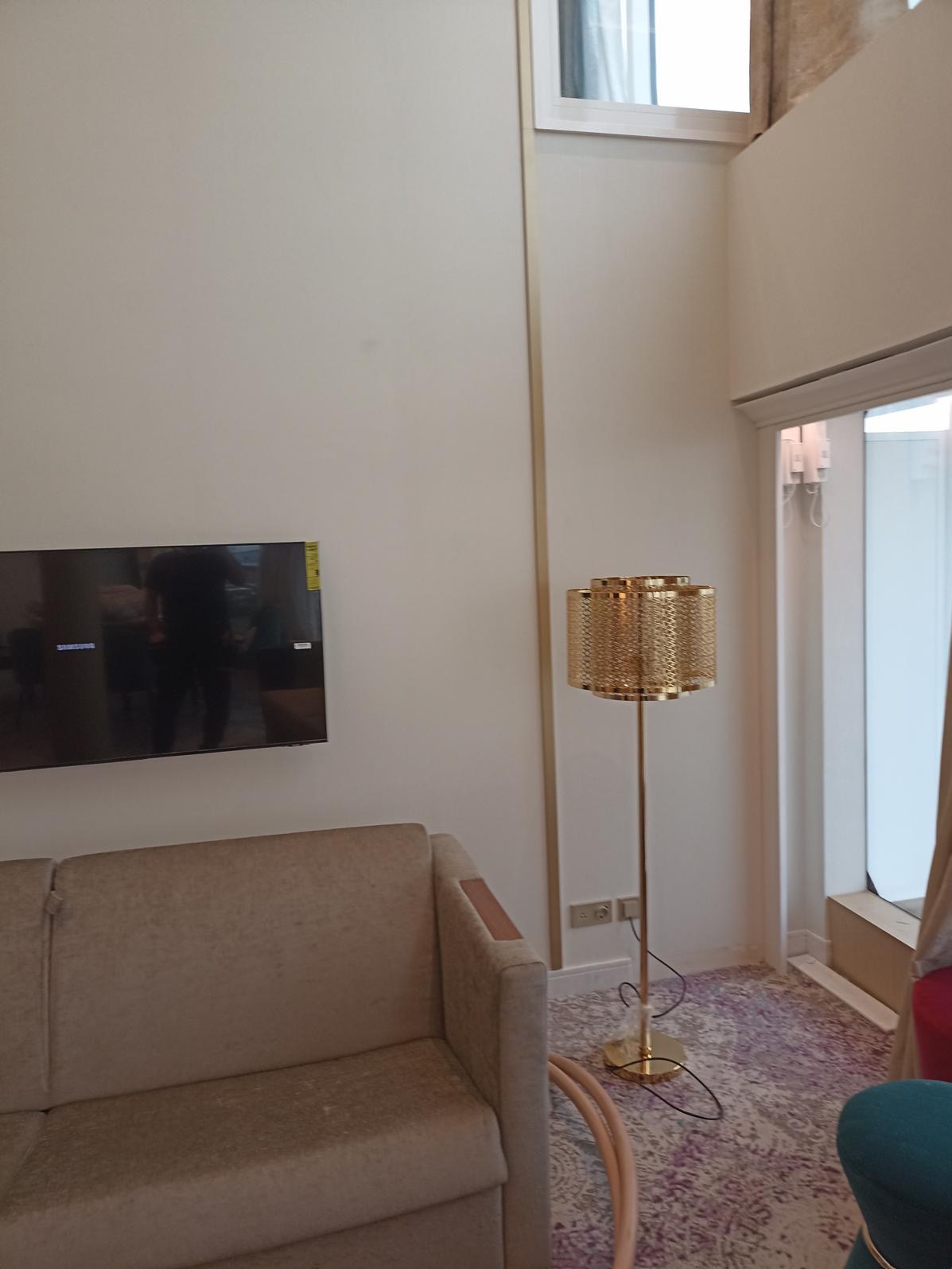 Jasne wnętrze z beżową sofą, telewizorem na ścianie i złotą lampą podłogową. Ściany w odcieniach bieli, subtelny wzór dywanu. Minimalistyczny design z akcentami złota.