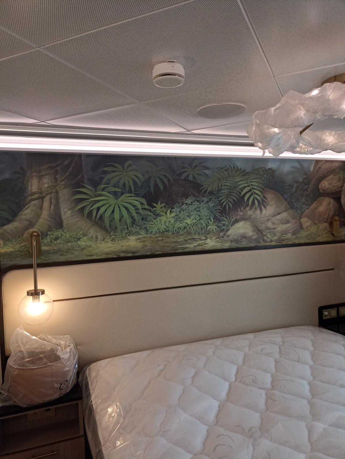 Pokój hotelowy z łóżkiem i malowidłem ściennym przedstawiającym dżunglę. Lampa nocna z kulistym kloszem. Sufit kasetonowy z czujnikiem dymu.