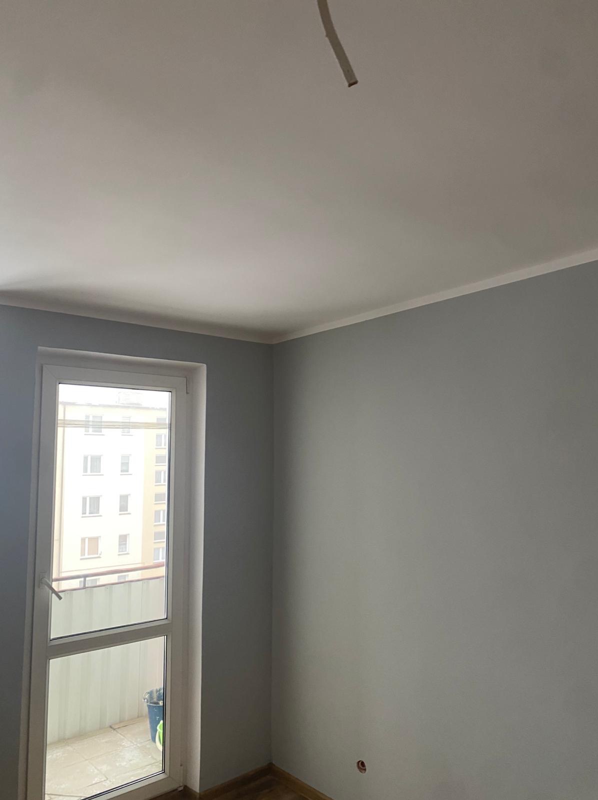 Jasnoszary pokój po malowaniu z białym sufitem i listwą. Widoczne okno balkonowe z widokiem na budynek. Fragment instalacji wystaje z sufitu.