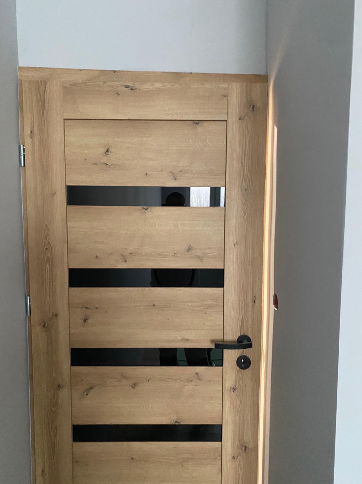 Zamknięte drzwi wewnętrzne z drewnianą okleiną i czarnymi, poziomymi przeszkleniami, widoczne na tle jasnej ściany. Czarna klamka po prawej stronie.