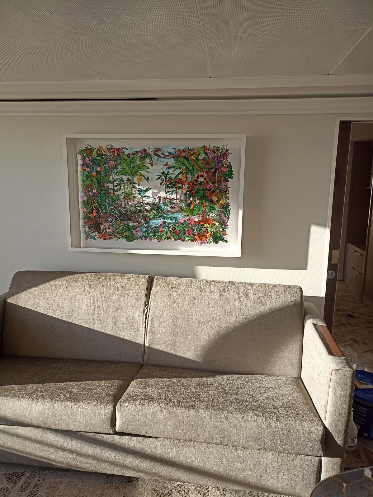 Salon z beżową sofą pod ścianą, na której wisi oprawiony w ramę obraz z motywem tropikalnym, oświetlony popołudniowym słońcem wpadającym przez okno.