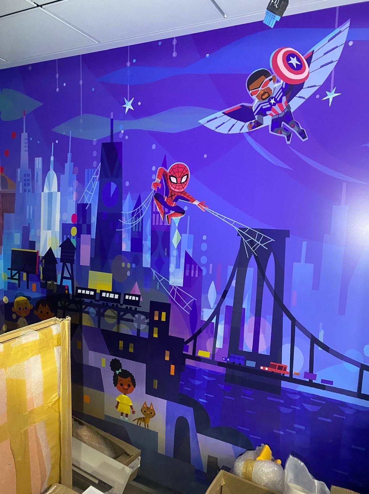 Kolorowa grafika ścienna w pokoju dziecięcym: Spiderman, Falcon, Brooklyn Bridge i panorama miasta w fioletowo-niebieskiej tonacji. W tle widoczne kartony i elementy wyposażenia.