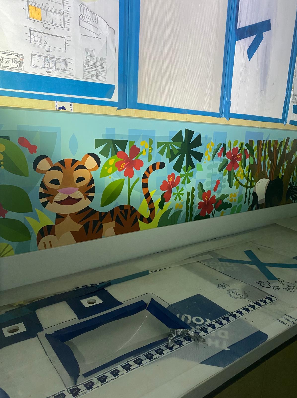 Kolorowa dekoracja ścienna z motywem dżungli i zwierząt (tygrys, panda) w pokoju dziecięcym, zabezpieczona taśmą malarską, z widocznymi szablonami na pierwszym planie.