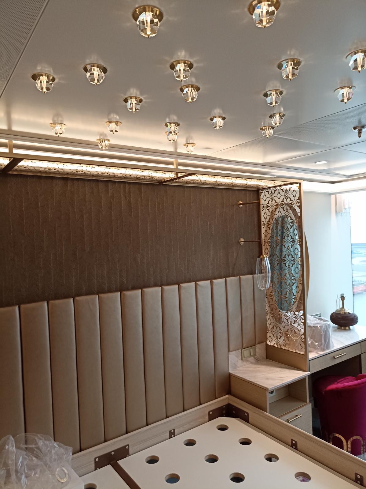 Luksusowa sypialnia w trakcie wykończenia: tapicerowany panel ścienny, dekoracyjne oświetlenie sufitowe, toaletka z lustrem w ozdobnej ramie. Widoczne elementy konstrukcyjne łóżka.
