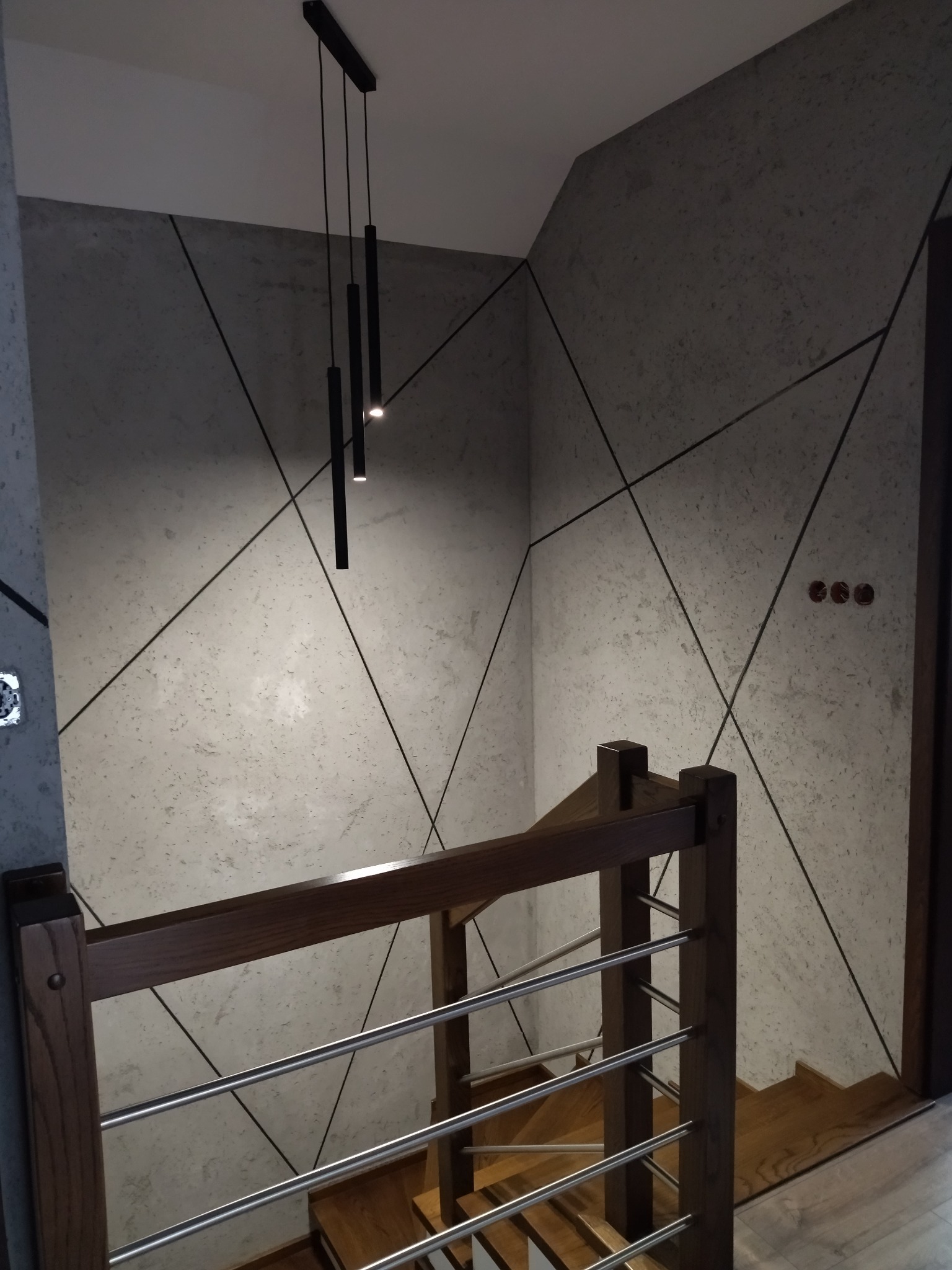 Nowoczesne wykończenie klatki schodowej z dekoracyjną szarą ścianą w geometryczne wzory, oświetloną wiszącą lampą. Widok z góry na drewniane schody i balustradę.