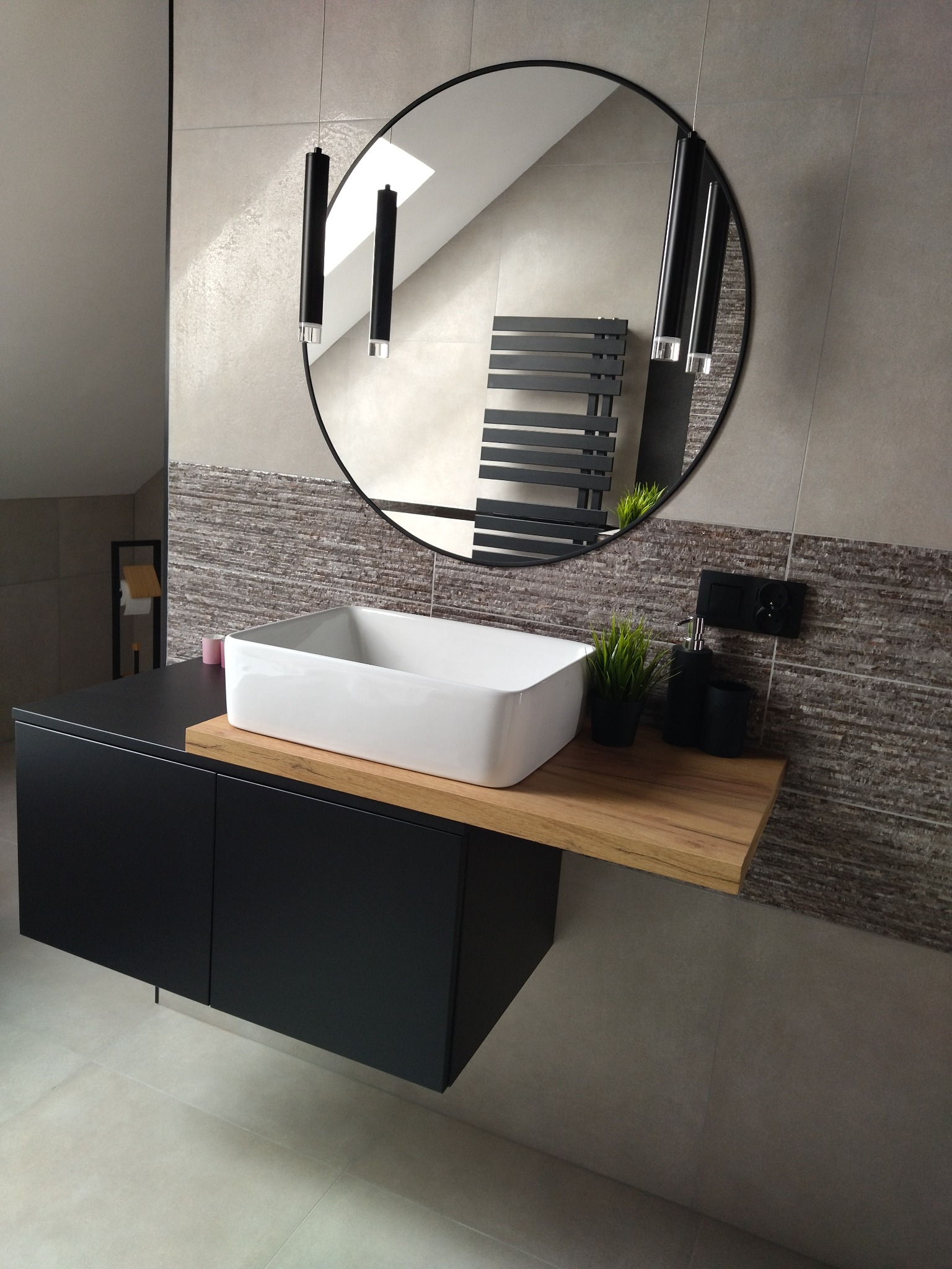 Nowoczesna łazienka z umywalką nablatową, czarną szafką i okrągłym lustrem. Na ścianie dekoracyjne płytki imitujące kamień. Minimalistyczne oświetlenie.