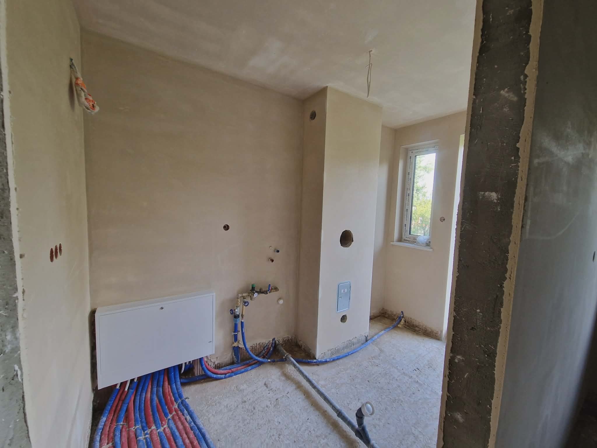 Wnętrze po tynkowaniu maszynowym: ściany w jasnym odcieniu beżu, widoczne instalacje wodne i ogrzewania podłogowego, okno z widokiem na zieleń. Surowy stan wykończenia.