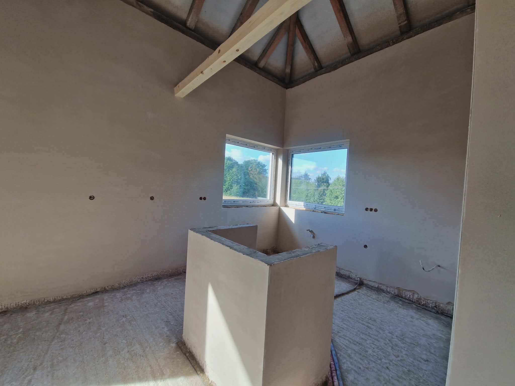 Wnętrze po tynkowaniu ścian i wylewce podłogi, widoczne okna z zielenią na zewnątrz i drewniana konstrukcja dachu. Ściany gładkie, przygotowane do dalszych prac.