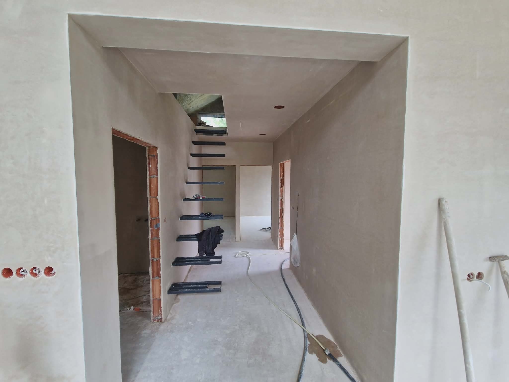 Wnętrze domu w trakcie prac wykończeniowych: ściany pokryte świeżym tynkiem, widoczne schody z metalowymi stopniami i przygotowane otwory pod gniazdka elektryczne.