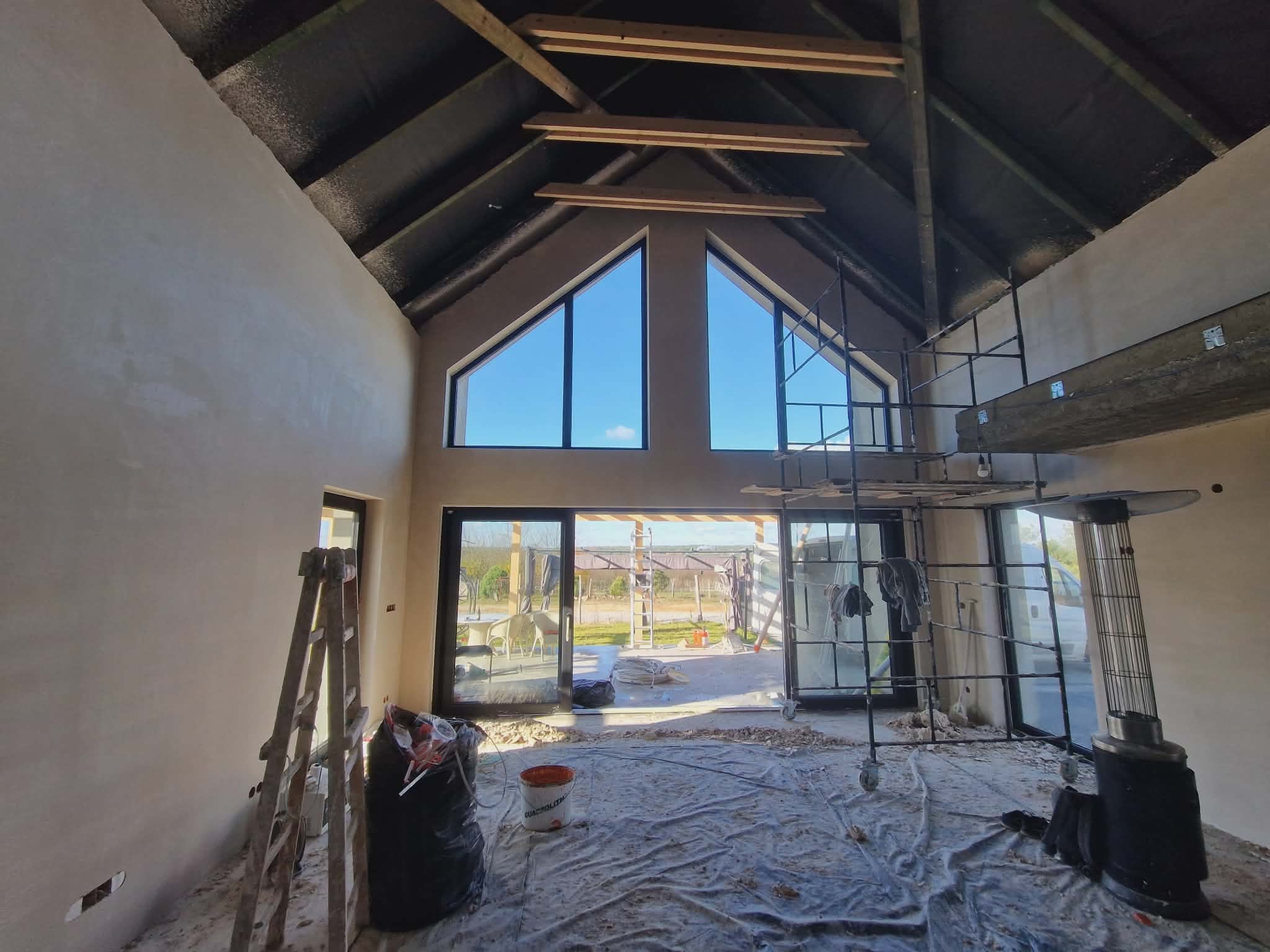 Wnętrze domu w trakcie prac wykończeniowych: ściany po tynkowaniu, widoczne rusztowania, okna trójkątne, przygotowane do dalszych prac. Podłoga zabezpieczona folią.