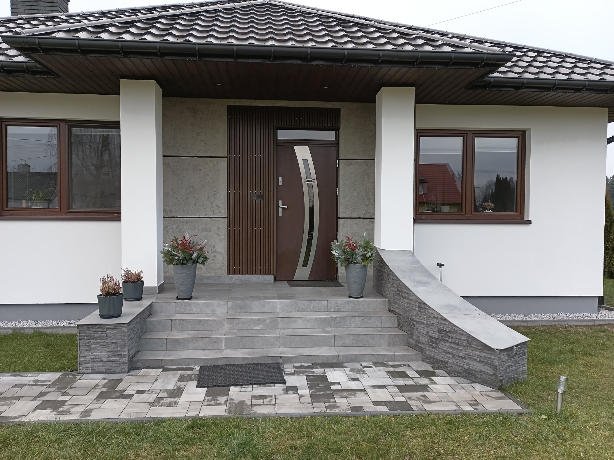 Elewacja domu jednorodzinnego z wejściem wyłożonym szarą kostką brukową i schodami. Widoczne okna z brązowymi ramami i dekoracyjne donice z roślinami.