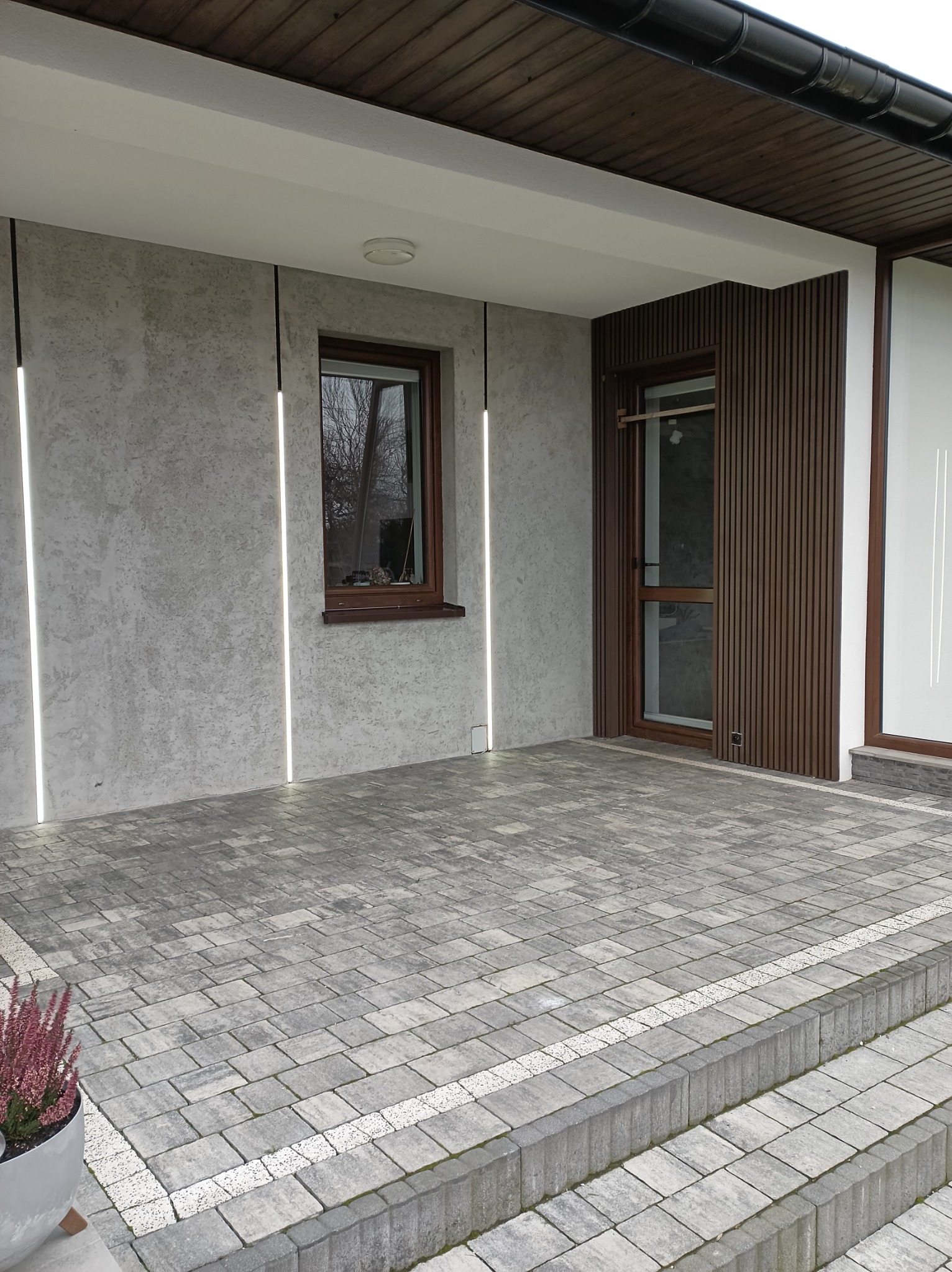 Wykończony fragment elewacji domu z szarą fakturą tynku, oświetleniem liniowym i drewnianą okładziną. Ułożona kostka brukowa na podjeździe.