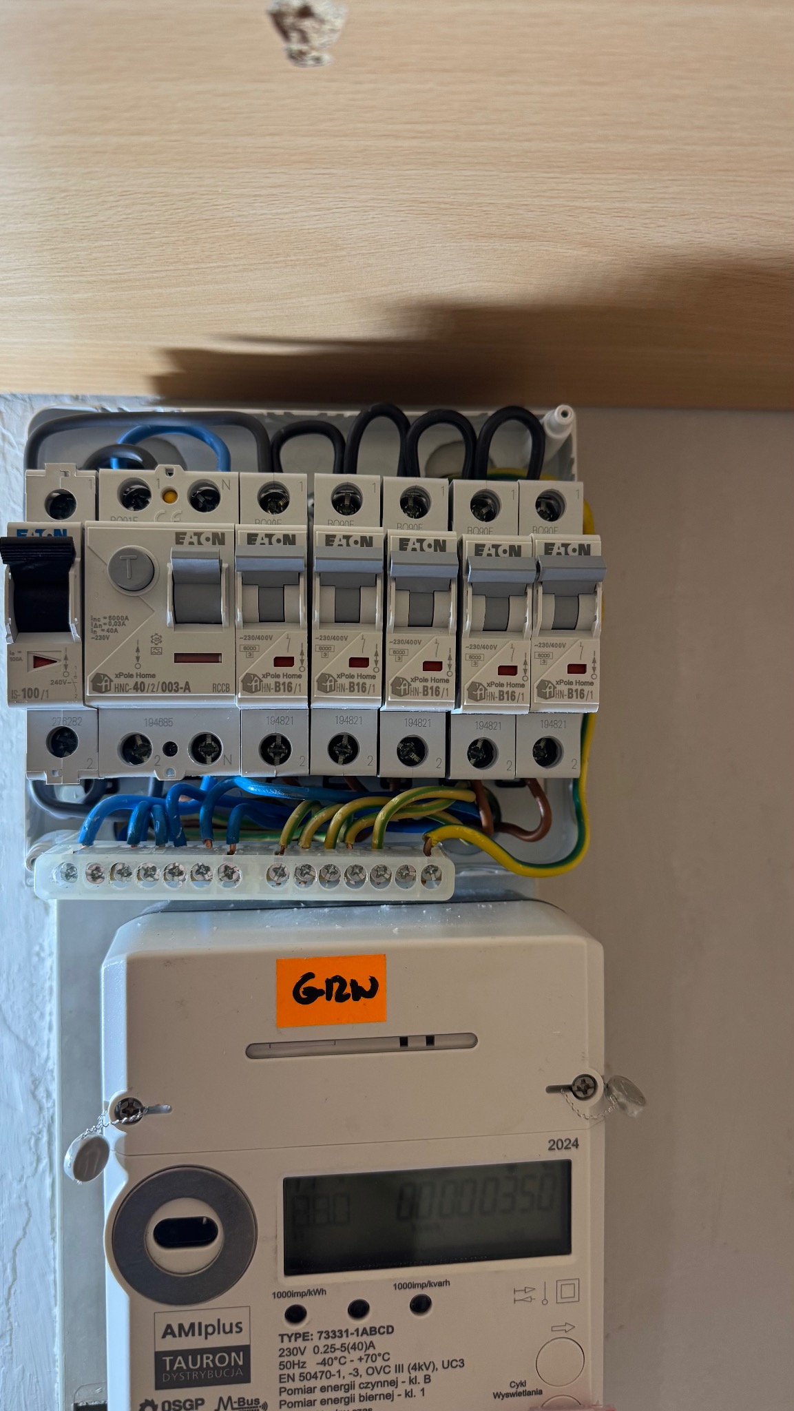 Rozdzielnica elektryczna z zamontowanymi bezpiecznikami Eaton i licznikiem Tauron, widoczne przewody i oznaczenia. Instalacja wewnątrz budynku, oświetlenie dzienne.