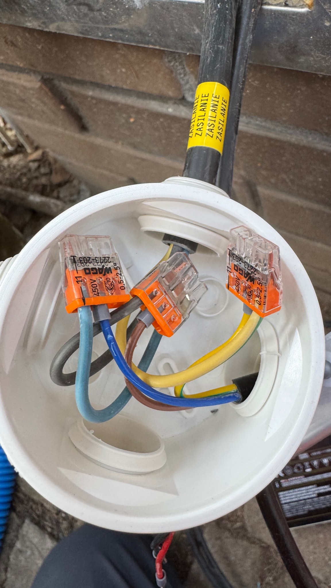 Biała puszka elektryczna z trzema złączkami WAGO, widoczne przewody w różnych kolorach. Żółta etykieta z napisem 'ZASILANIE' na czarnym kablu.