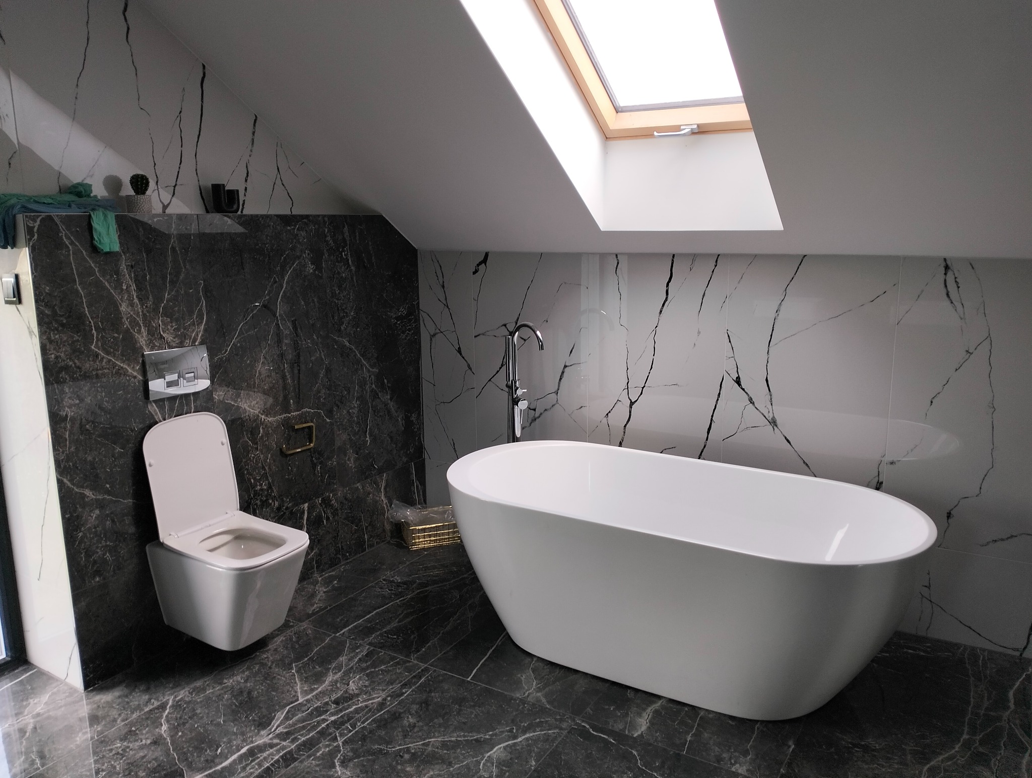 Nowoczesna łazienka na poddaszu z czarno-białymi płytkami imitującymi marmur, wolnostojącą wanną i podwieszaną toaletą. Okno dachowe doświetla wnętrze.