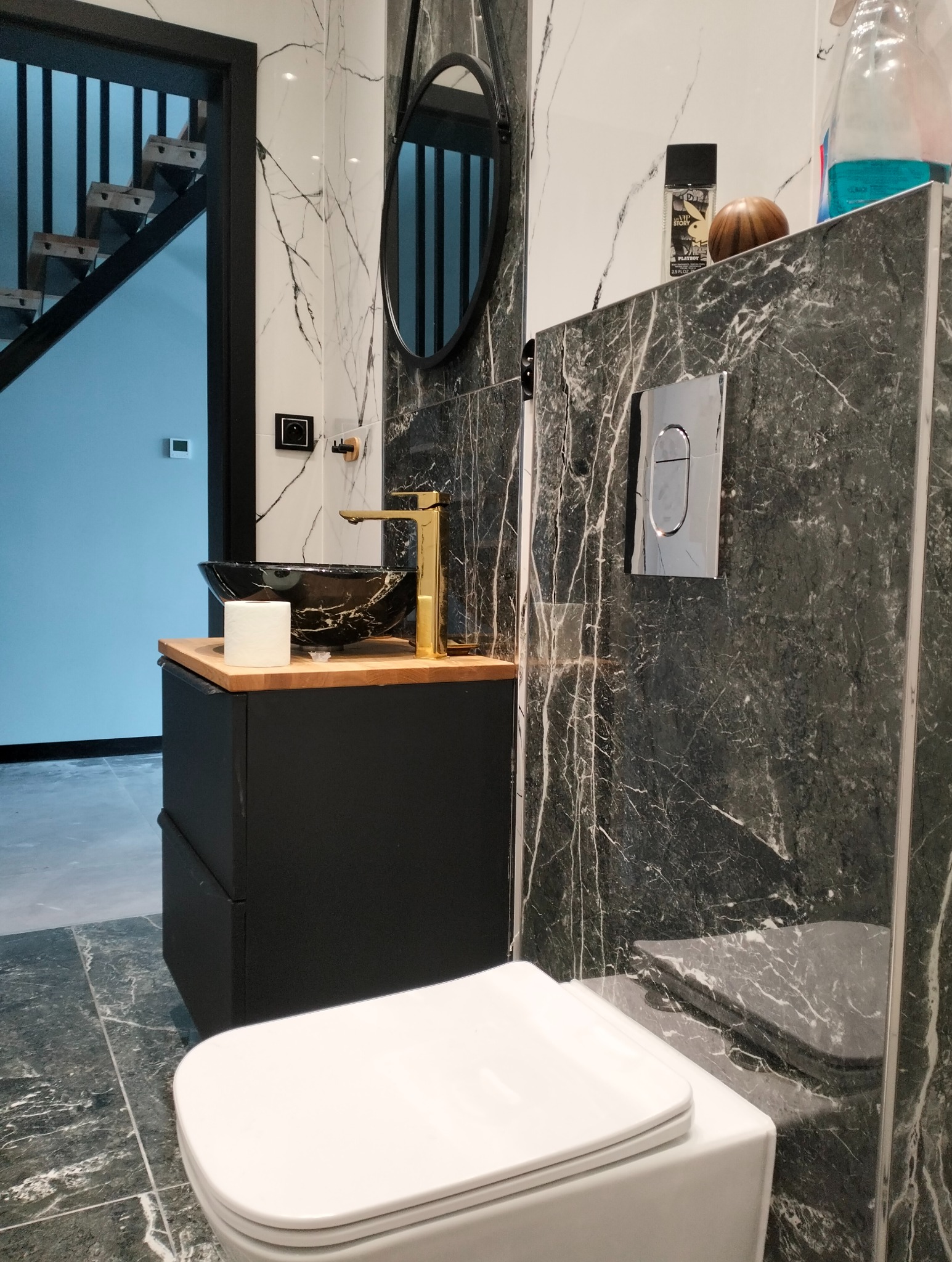 Nowoczesna łazienka z marmurowymi płytkami, czarną szafką z umywalką w stylu glamour i złotą armaturą. Widoczne schody w tle.