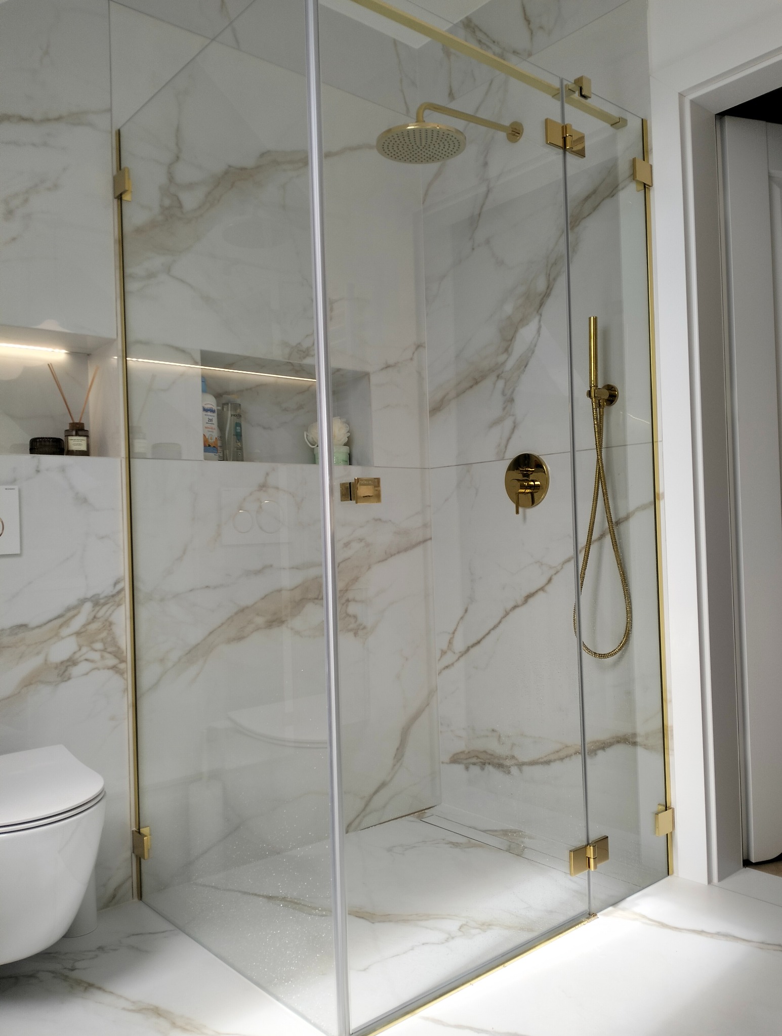 Nowoczesna kabina prysznicowa z elementami w kolorze złota, marmurowe płytki na ścianach i podłodze, widoczna toaleta w tle. Eleganckie wykończenie łazienki.