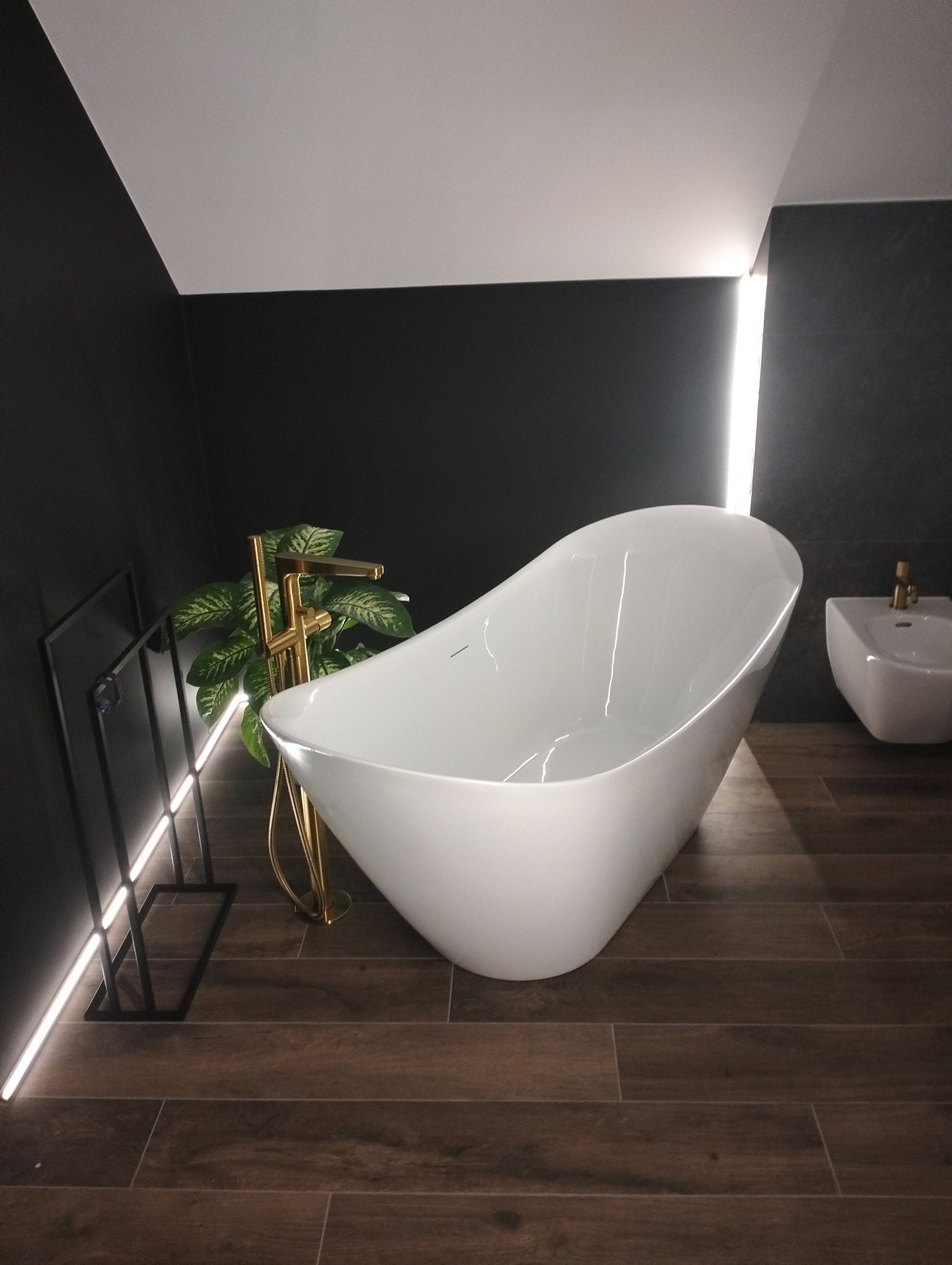 Nowoczesna łazienka z wolnostojącą białą wanną, złotą armaturą i drewnopodobną podłogą. Minimalistyczny design z akcentem roślinnym i podświetleniem LED.