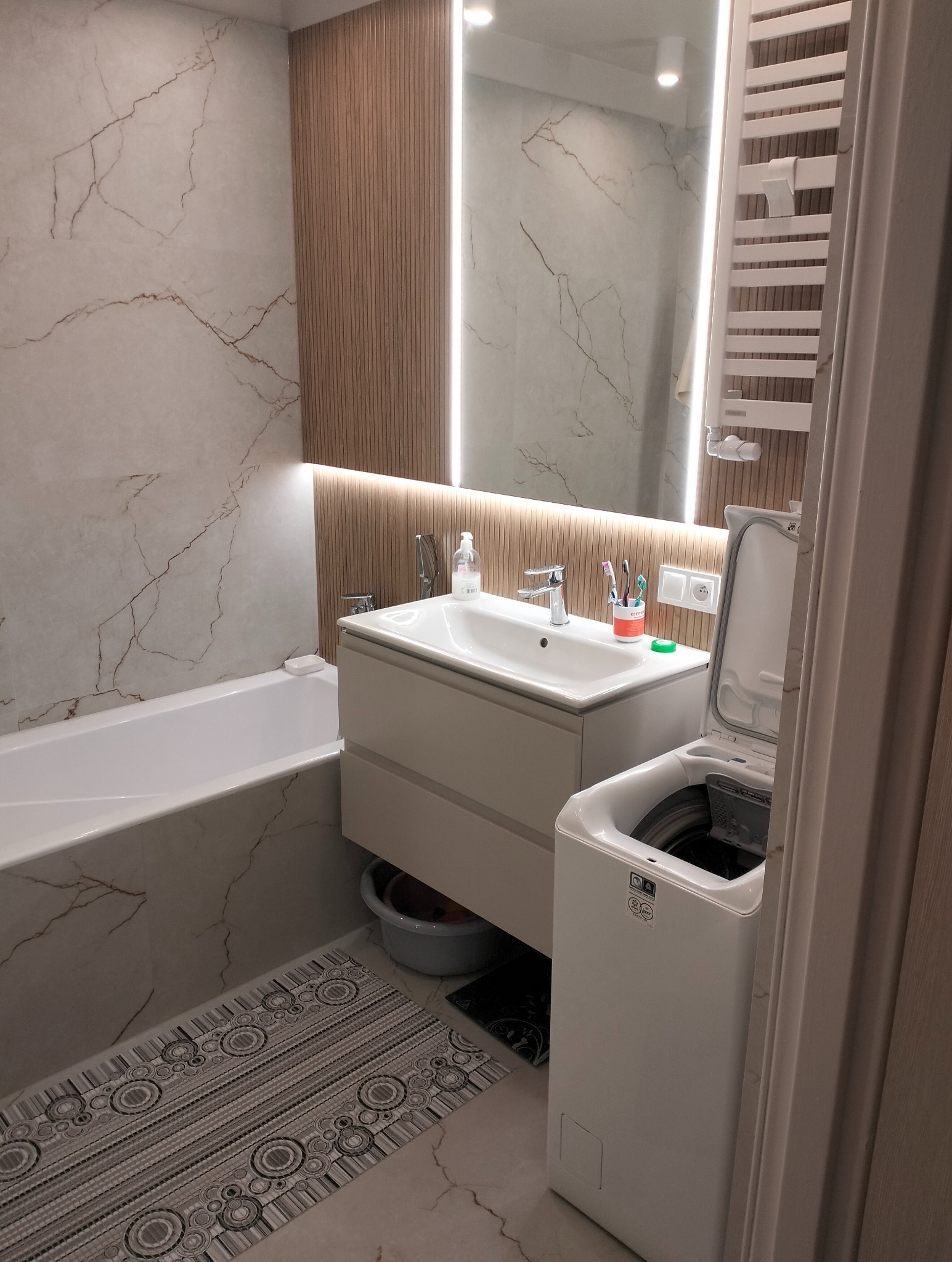 Nowoczesna łazienka z marmurowymi płytkami, drewnianymi panelami i podświetlanym lustrem. Umywalka z szafką, wanna i pralka tworzą spójną całość.