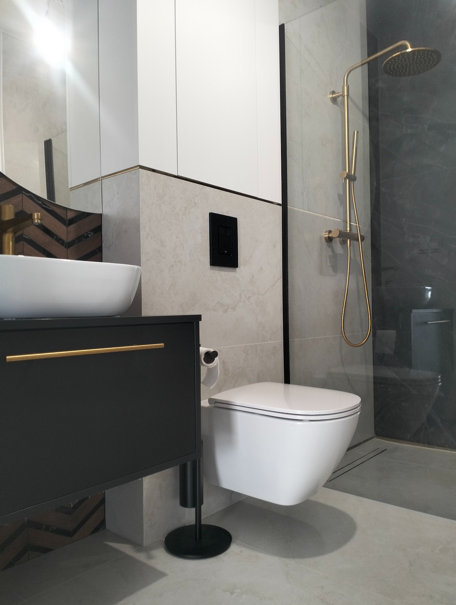 Nowoczesna łazienka z wiszącą toaletą, umywalką na szafce z złotymi uchwytami i złotą armaturą prysznicową. Płytki w odcieniach szarości i beżu, geometryczny wzór za umywalką.