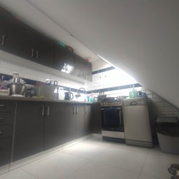 usługi ogólnobudowlane - Kuchnia w szarościach z białymi płytkami w układzie cegiełki, z czarnym pasem. Uwagę zwraca skośny sufit i podświetlenie szafek. Widoczny sprzęt AGD.