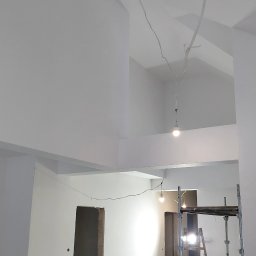 Działalność ogolnobudowlana - Białe, świeżo pomalowane wnętrze z widocznymi detalami architektonicznymi i rusztowaniem. W tle surowe otwory drzwiowe. Oświetlenie tymczasowe.