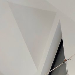 Działalność ogolnobudowlana - Biały sufit z geometrycznymi załamaniami i oknem w tle, oświetlony żarówką na kablu. Minimalistyczne wykończenie wnętrza, nowoczesny design.