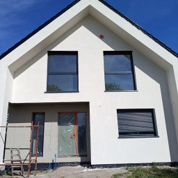 Działalność ogolnobudowlana - Front nowoczesnego domu z jasną elewacją, czarnym dachem i dużymi oknami. Widoczne rusztowanie, folia kubełkowa przy fundamencie oraz drzwi zabezpieczone folią, pod błękitnym niebem.