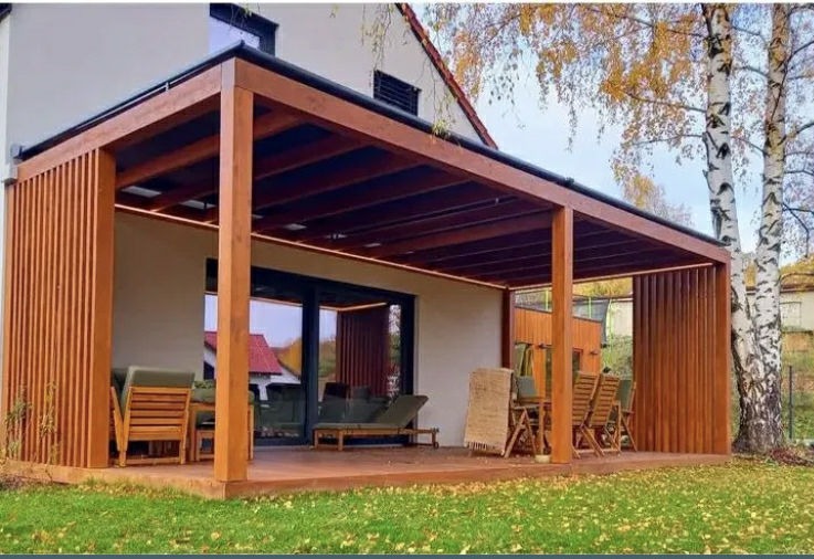 Drewniana pergola z oświetleniem LED, przylegająca do domu. Widoczne meble ogrodowe i drzewo z żółtymi liśćmi. Ujęcie z poziomu trawnika.