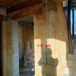 WOJ-TECH Wojciech Świderski - Surowy stan budynku w trakcie prac instalacyjnych: widoczne przewody elektryczne prowadzone po ścianach z czerwonymi puszkami, sprzęt budowlany w tle, cień osoby na ścianie.