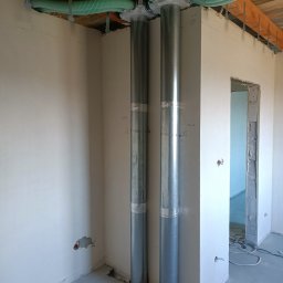 WOJ-TECH Wojciech Świderski - Dwie pionowe, metalowe rury wentylacyjne z zielonymi, elastycznymi przewodami pod sufitem w surowym wnętrzu. Widoczne ślady po instalacji i niedokończone ściany.