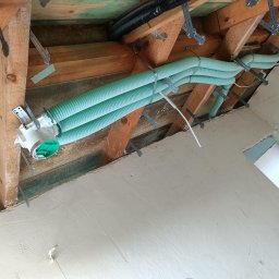 WOJ-TECH Wojciech Świderski - Zielone rury wentylacyjne zamocowane do drewnianej konstrukcji dachu na poddaszu, widoczne elementy instalacji i fragment otynkowanej ściany.