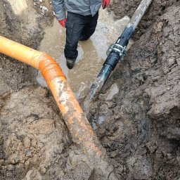 WOJ-TECH Wojciech Świderski - Połączenie rur w wykopie wypełnionym wodą. Widoczne pomarańczowe rury kanalizacyjne i czarna rura z metalowym łącznikiem. Osoba w roboczym ubraniu stoi w wodzie.