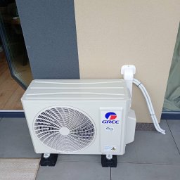 WOJ-TECH Wojciech Świderski - Biała jednostka zewnętrzna klimatyzacji Gree zamontowana na balkonie, z widocznymi przewodami i elementami mocującymi. Tło stanowi elewacja budynku w dwóch odcieniach.