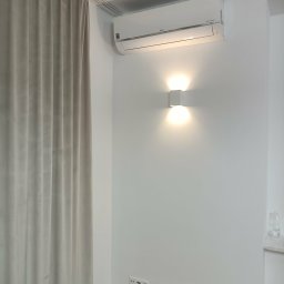 WOJ-TECH Wojciech Świderski - Biały klimatyzator LG na ścianie nad kwadratową lampą ścienną, obok zasłony. Widoczne gniazdka elektryczne. Minimalistyczne wnętrze.